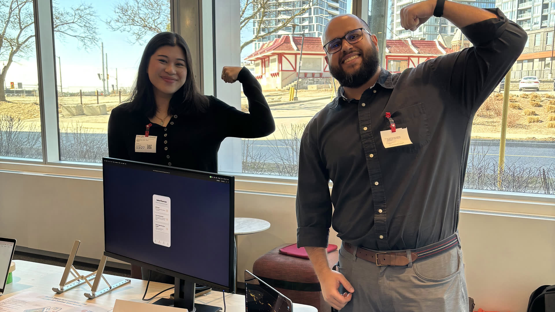 Two students flexing their arms beside the monitor showing their MuscleMemory prototype at their demo station.
